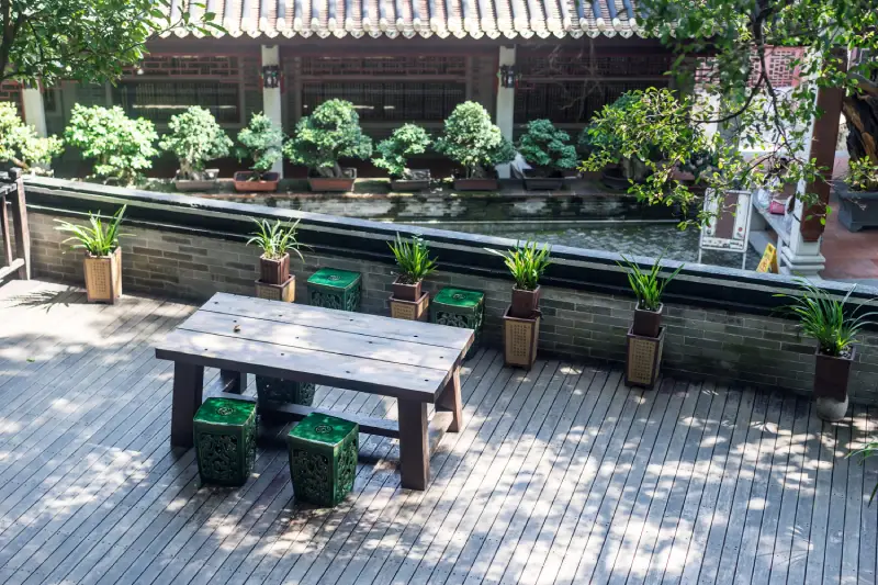 A wooden table with green stools sits on a deck surrounded by potted plants, overlooking a tranquil pond and traditional architecture.